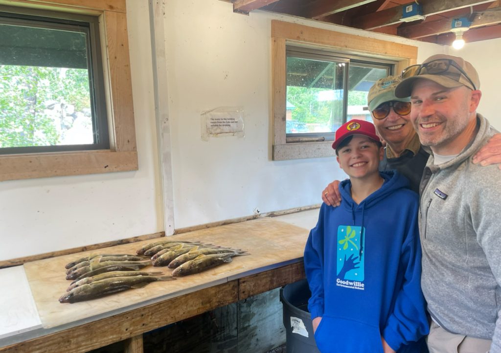 A smiling trio poses in a fishing cabin with their catch on Lake Vermillion. A boy in a blue hoodie and red hat stands in the front, while two adults in sunglasses stand behind. Several fish lie on a table before them. The wooden windows of the cabin allow light to stream into this 2024 fishing report snapshot.