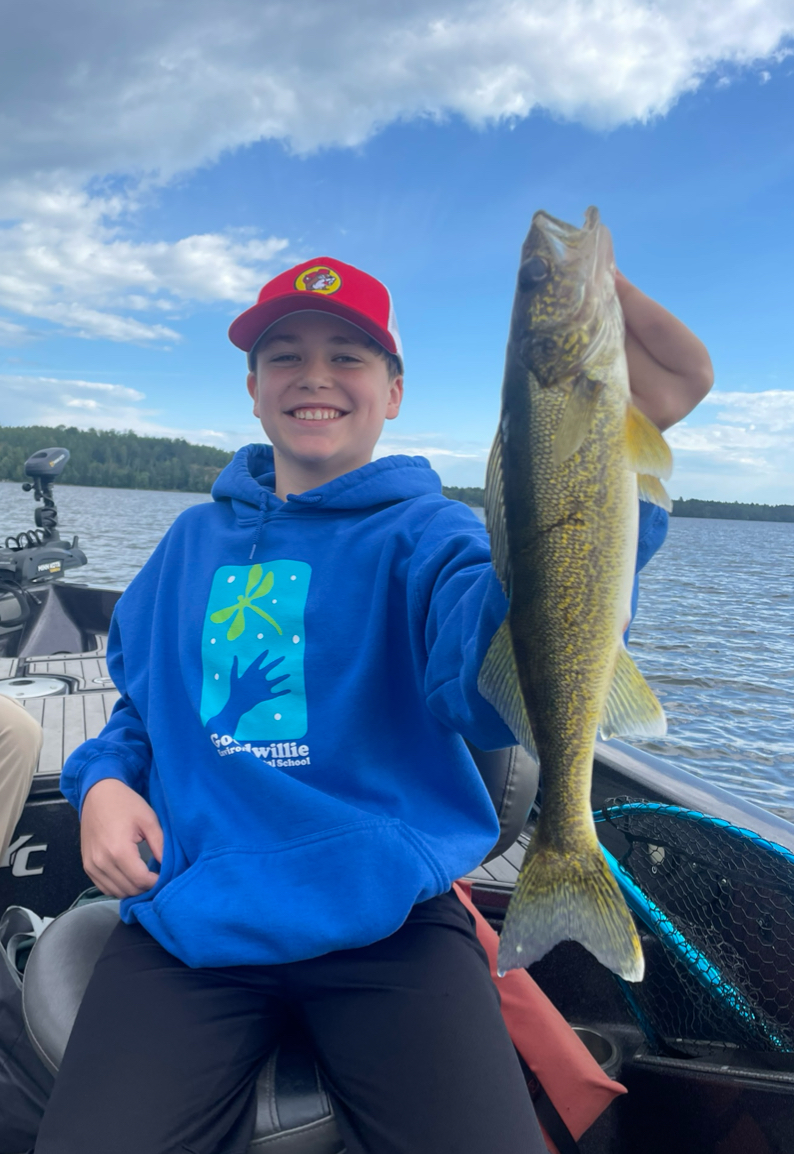 A young person is smiling while holding a large fish on a boat in Lake Vermillion. They are wearing a red cap and a blue hoodie with a design on it. The background features the serene body of water and a partly cloudy sky, making for an excellent 2024 fishing report photo.