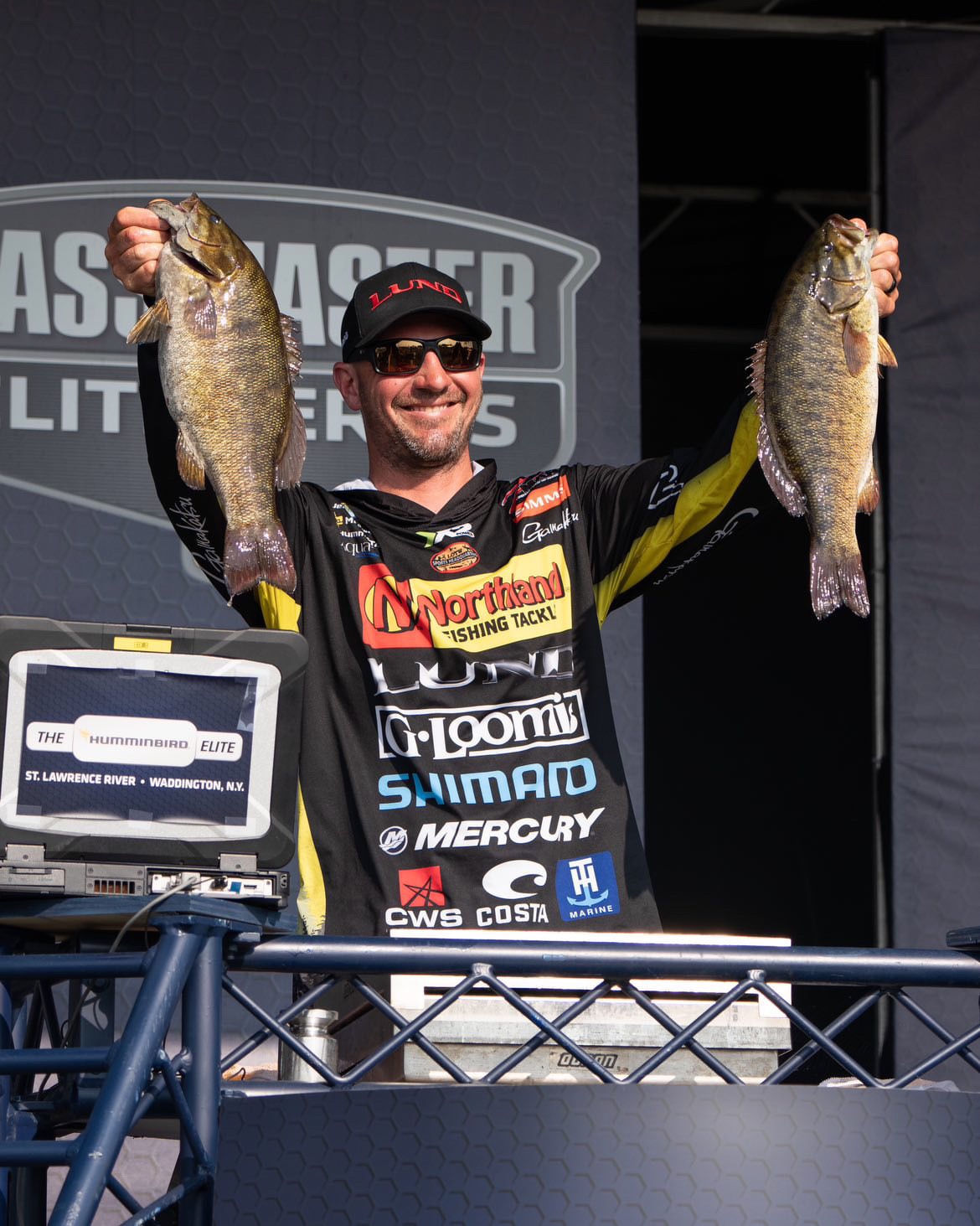 A person in a black and yellow fishing jacket and cap proudly holds up two fish on stage, set against Ultimate Fishing competition branding. A cooler and electronic device sit on the table before them, capturing this electrifying moment of their fishing adventure.