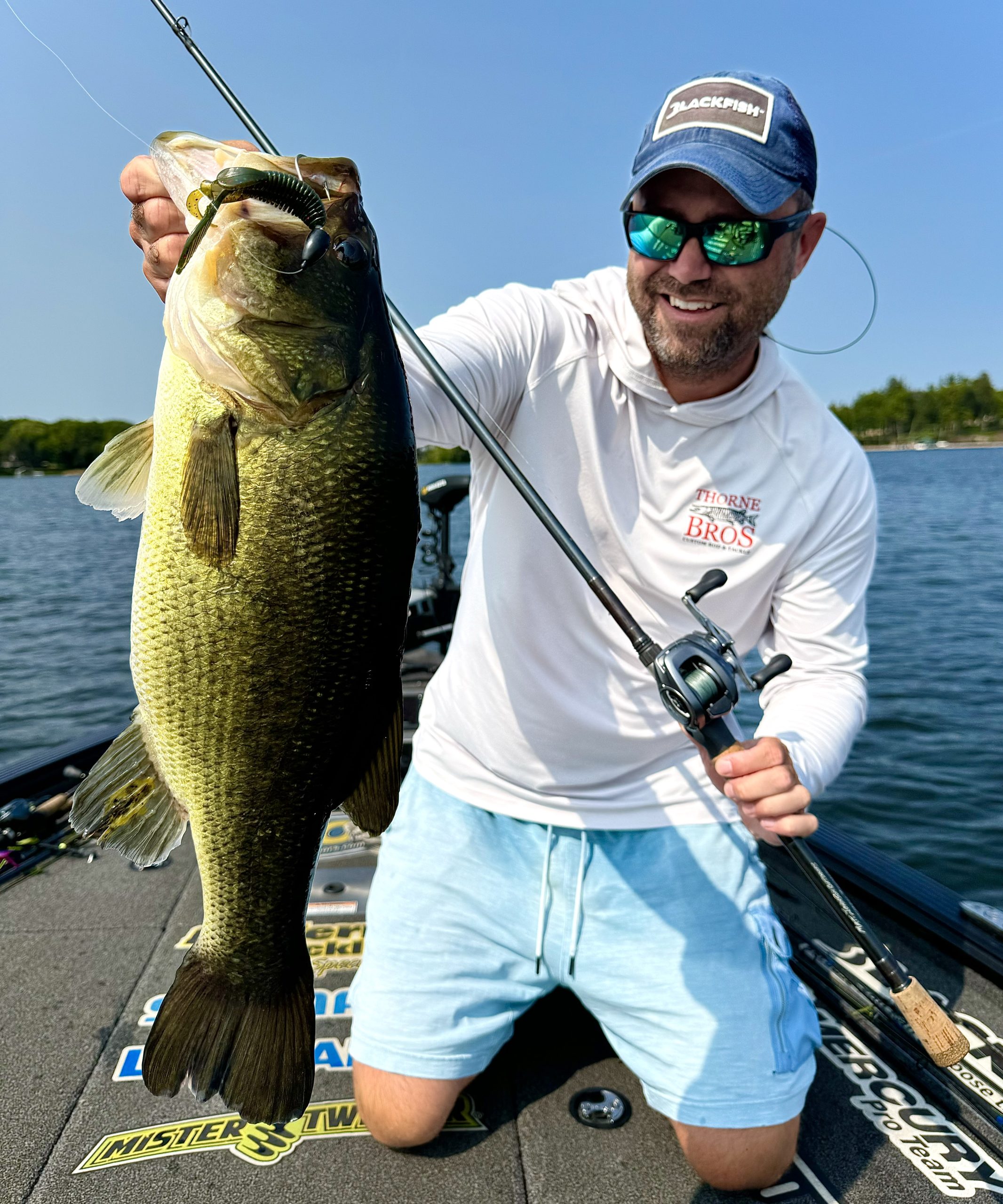 A person in a white shirt and blue shorts is kneeling on a boat, proudly holding their ultimate catch. They are wearing a cap, mirrored sunglasses, and smiling. The background showcases the serene body of water and blurred greenery, creating the perfect fishing adventure.