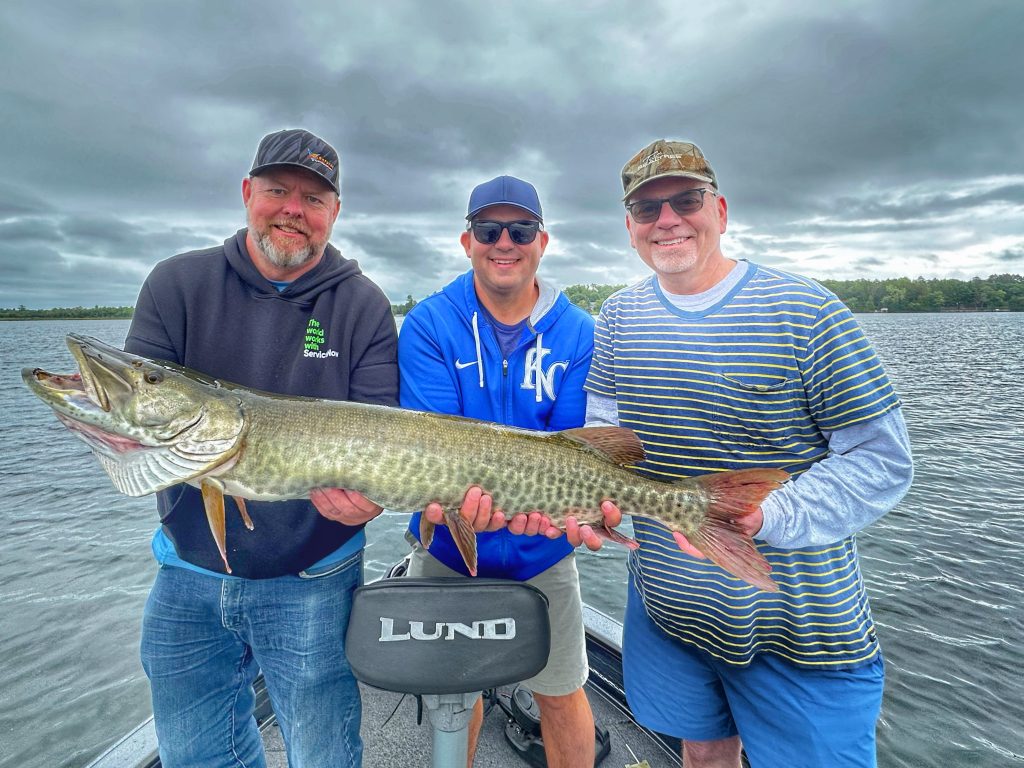 Three men stand on a boat at Leech Lake, holding a large fish and smiling at the camera. It's the perfect fishing adventure; the sky is overcast and the water calm, as they enjoy their triumphant catch in casual outdoor clothing.