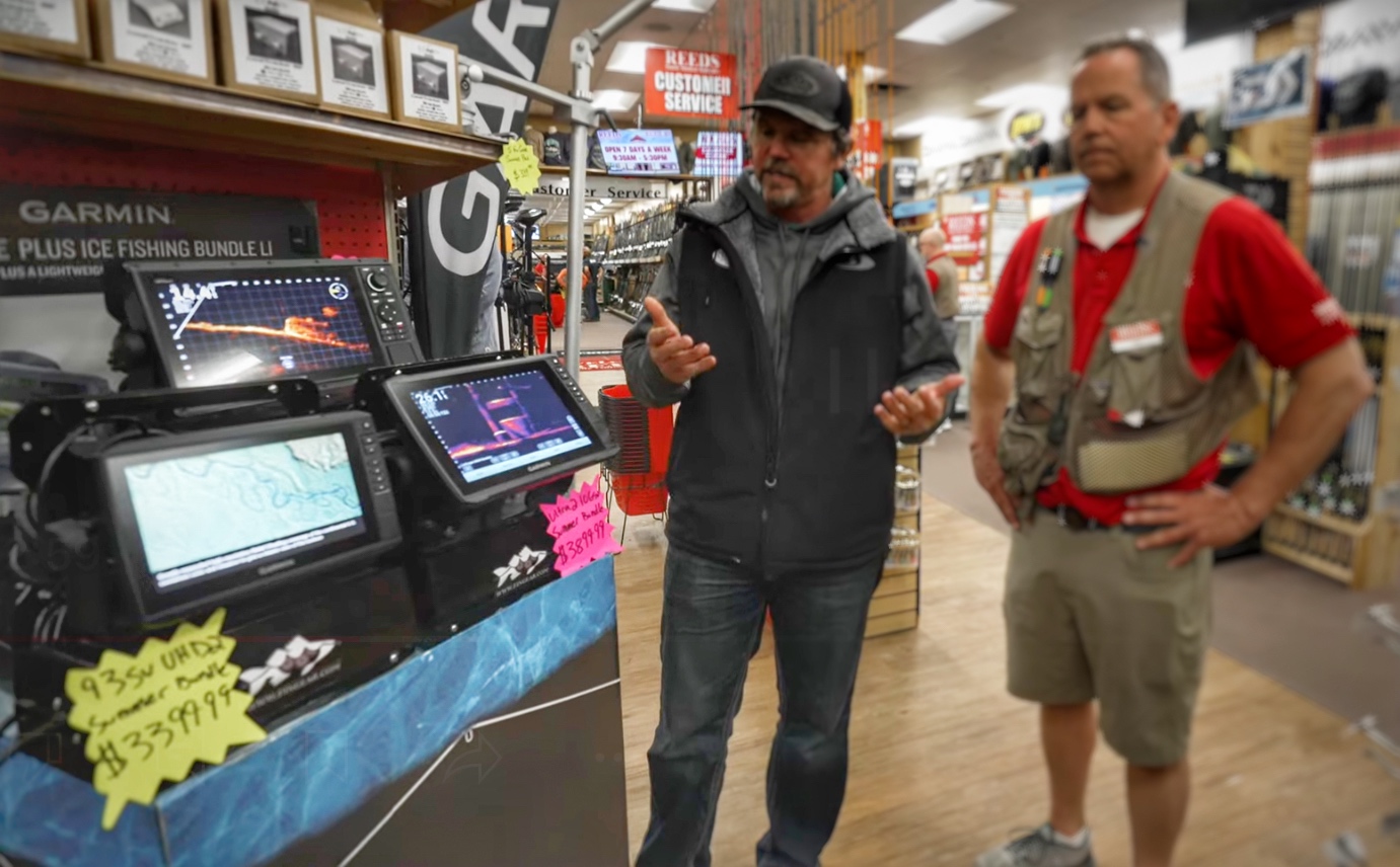 Two men in a store discuss cutting-edge ice fishing technology displayed on screens with product signs. One man shares expert tips while the other listens intently. The background features shelves brimming with equipment for walleye fishing adventures.