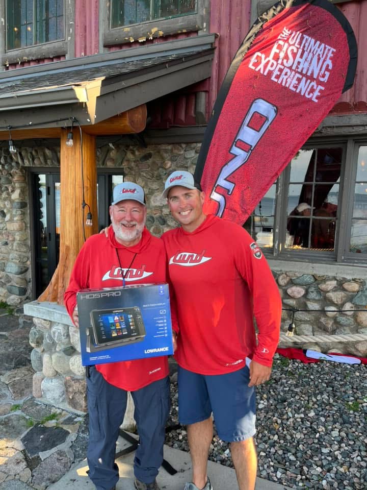 Two men in matching red lund shirts and hats smile while holding a fishing equipment box. They stand in front of a rustic building with a large banner that reads 