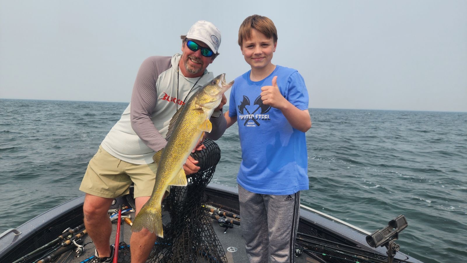 An adult and a child on a boat at Gull Lake proudly display the large fish they caught. The adult, in sunglasses, hat, long-sleeve shirt, and shorts, beams with pride. The child gives a thumbs up in a blue T-shirt and shorts as the expansive ocean stretches into the horizon behind them.