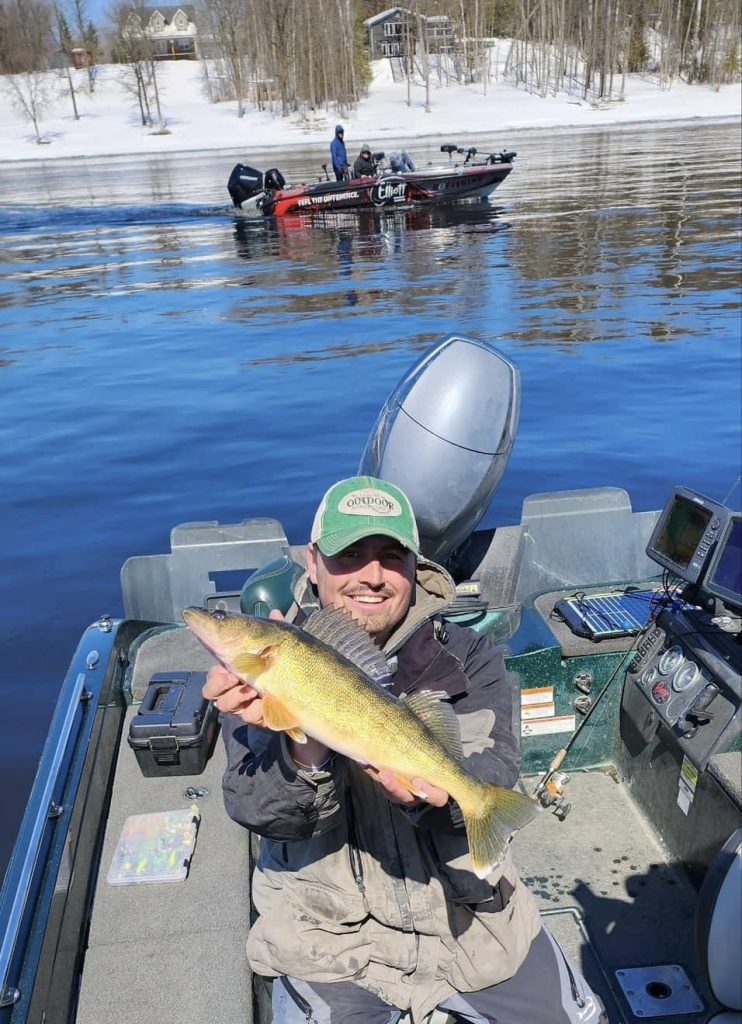 A person in a boat proudly holds a large fish, smiling at the camera during an early Walleye fishing adventure. The boat is equipped with fishing gear and floats on a calm, reflective body of open water. Another boat with two people is in the background against trees and a snowy shoreline.