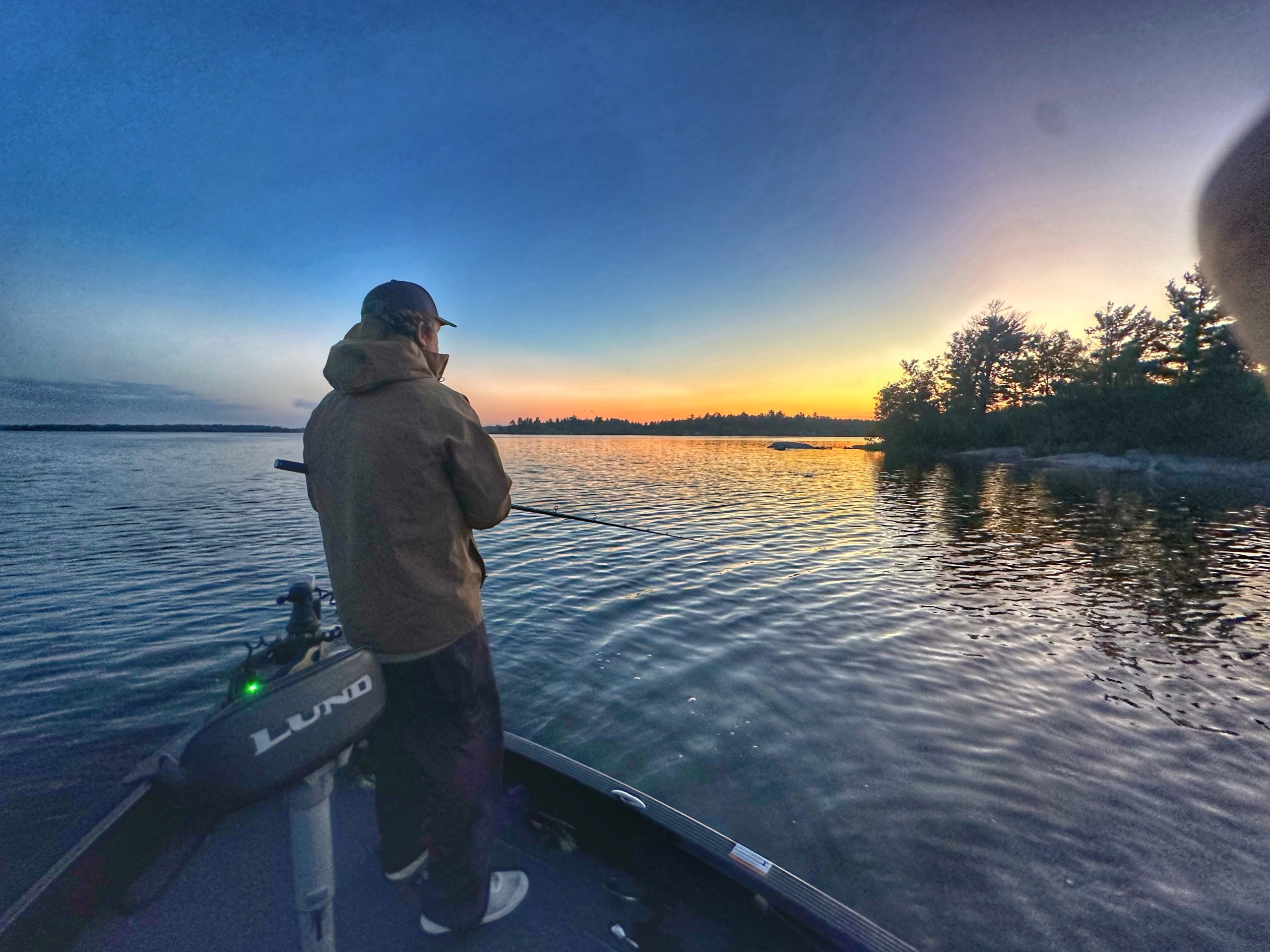A person in a jacket stands in a boat fishing at sunset on one of Minnesota's best fishing lakes. The sky is a gradient of blue to orange, with trees silhouetted in the distance, and the water mirrors the colorful sky.