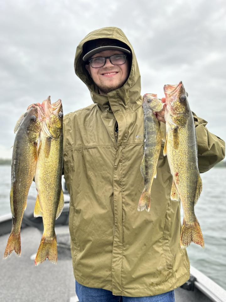A smiling fishing guide client stands on a boat at Gull Lake Minnesota, holding up four large walleye —two in each hand—with cloudy skies and water in the background, capturing the thrill of fishing.