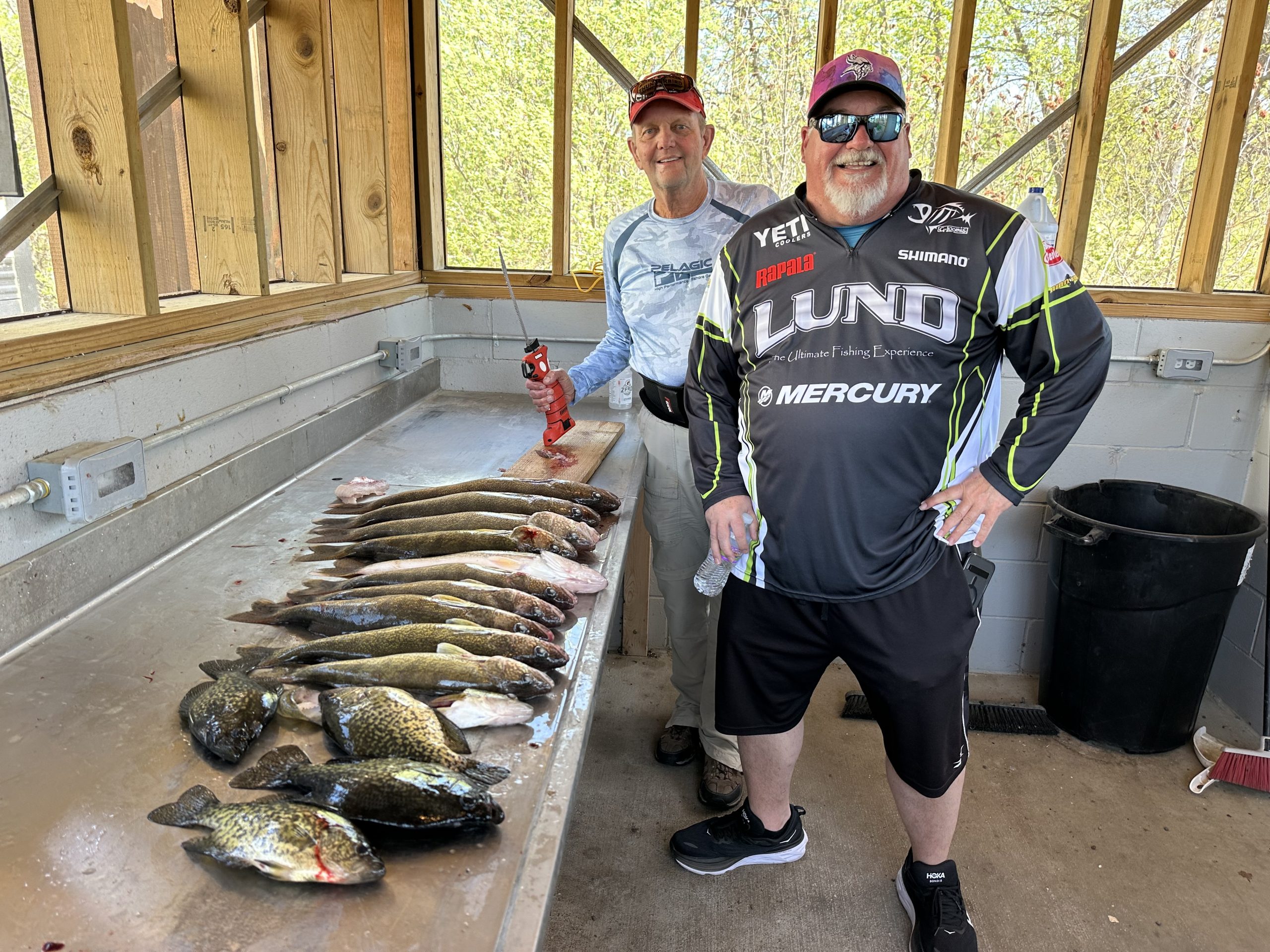 Two smiling men stand in a fish cleaning station; one, dressed as a fishing guide, cleans fish at the counter while the other poses in fishing gear. Several cleaned fish are laid out on the counter in front of them.