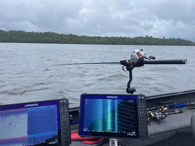 A fishing rod rests on the edge of a boat above choppy Lake Vermilion water, with two electronic fish finders displaying sonar images in the foreground. Trees line the distant shore under a cloudy sky—perfect for targeting walleyes.