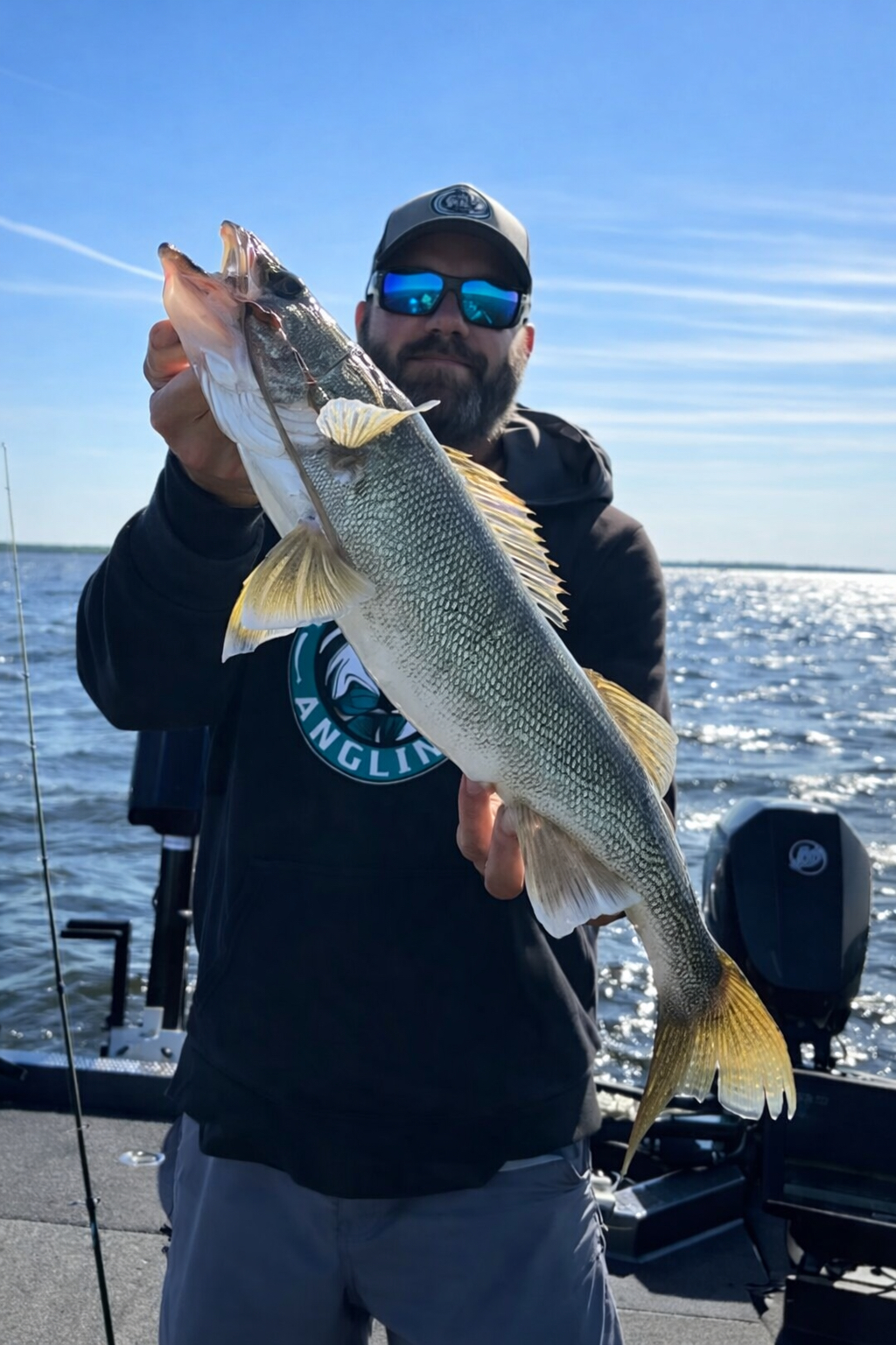 A man wearing sunglasses and a hoodie holds up a large fish on a boat with a lake and blue sky in the background.