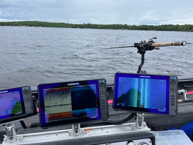 Three fish finder screens display underwater sonar images on a fishing boat facing a lake. A fishing rod is mounted on the boat’s edge, and electronics help anglers learn about the waters beneath a cloudy sky lined with distant trees.