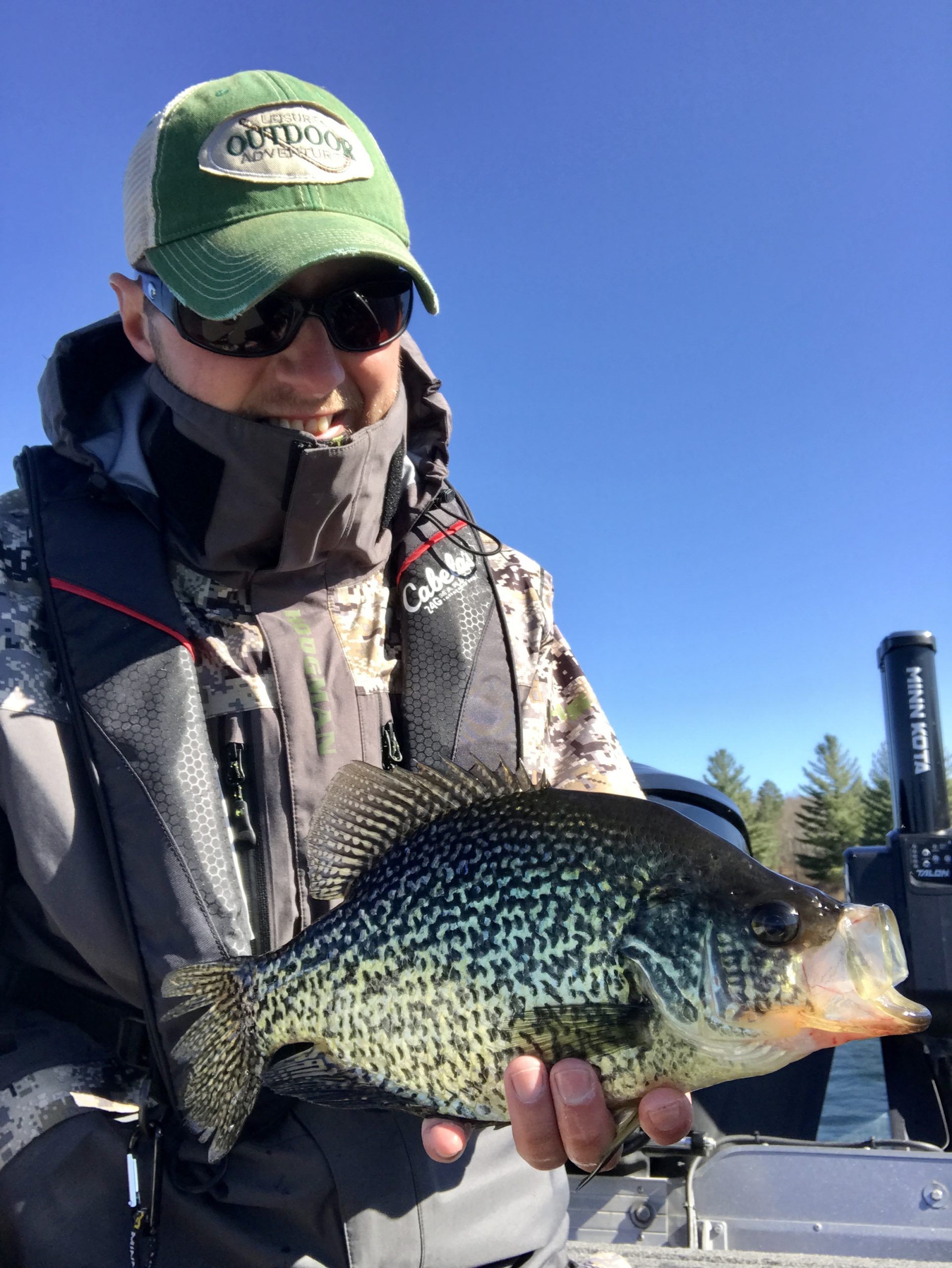 A person wearing sunglasses, a green cap, and a camo jacket smiles while holding a large crappie fish on a boat, with blue sky and trees in the background.