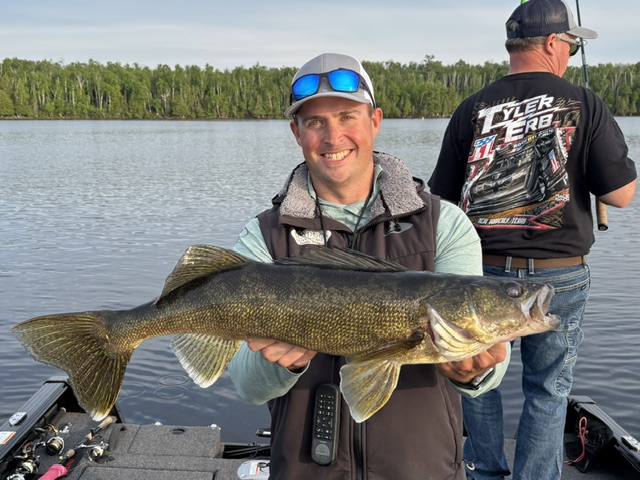 A smiling man in a cap and sunglasses holds a large fish on a boat in a lake, with trees in the background. Another man stands behind him, fishing and wearing a racing-themed shirt.